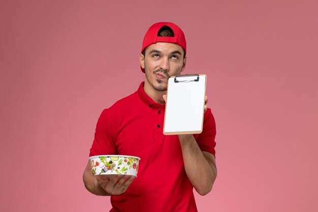 Giovane corriere maschio di vista frontale in capo uniforme rosso che tiene ciotola di consegna rotonda e blocco note pensando su sfondo rosa.