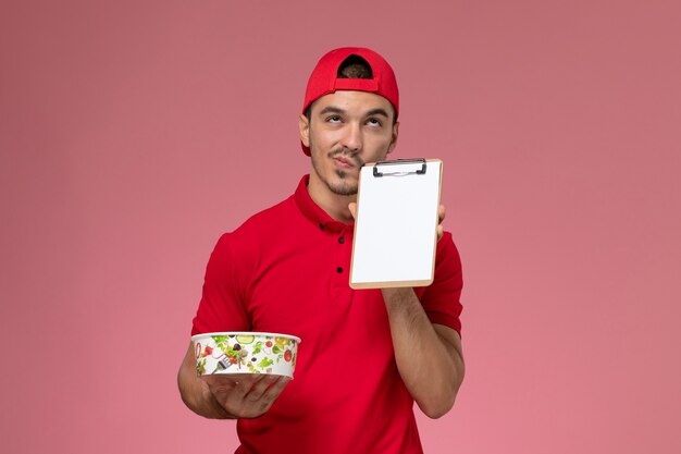 Front view young male courier in red uniform cape holding round delivery bowl and notepad thinking on pink background.
