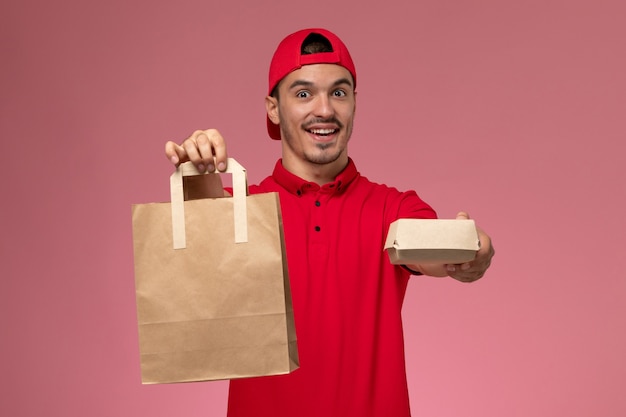 Foto gratuita corriere maschio giovane vista frontale in mantello rosso uniforme che tiene due diversi pacchetti di cibo sullo sfondo rosa.