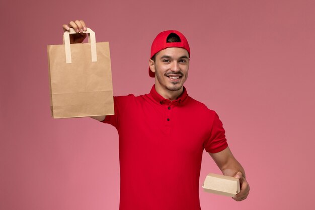 ピンクの背景に2つの異なる食品パッケージを保持している赤い制服の岬の正面図若い男性の宅配便。