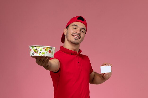 Молодой курьер-мужчина в красной форме, держащий белую пластиковую карту и миску для доставки на светло-розовом фоне, вид спереди.