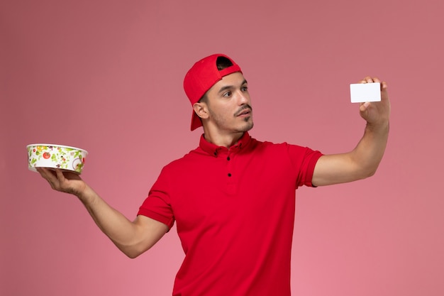 Front view young male courier in red uniform cape holding white plastic card and delivery bowl on light-pink background.