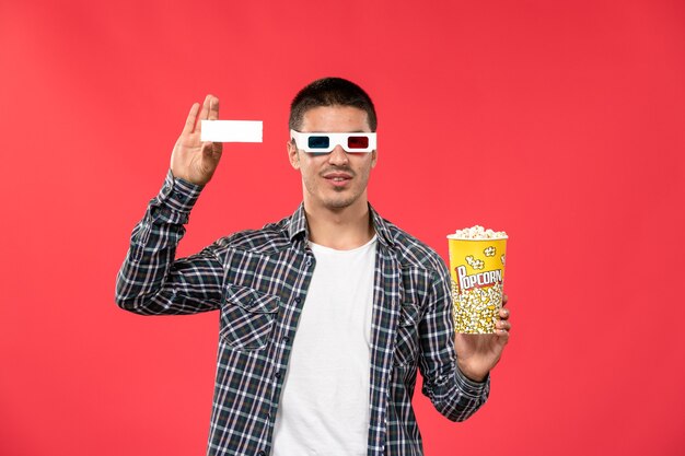 Front view young male in -d sunglasses holding popcorn package and ticket on light red wall cinema movies theater film male