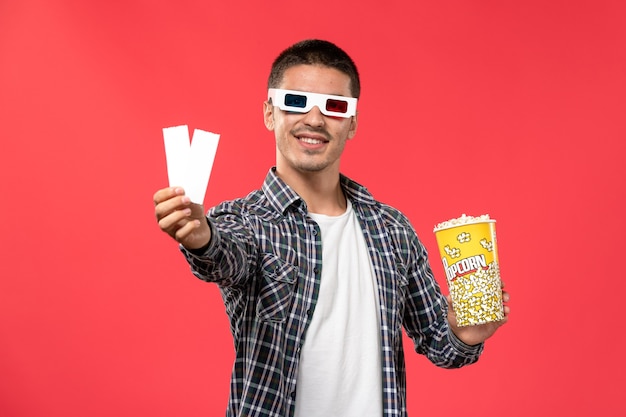 Вид спереди молодой мужчина в солнцезащитных очках -d с пакетом попкорна и билетами на светло-красной стене кинотеатр кинотеатр фильм мужчина
