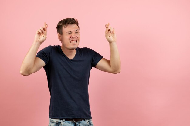 Front view young male in dark-blue shirt posing crossing his fingers on pink background