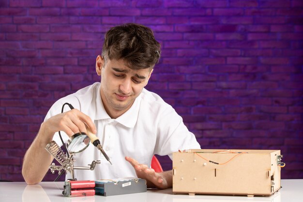 Front view young male behind desk trying to fix little construction layout on purple wall