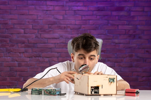 Front view young male behind desk trying to fix little house layout on purple wall