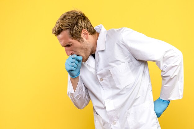 Вид спереди молодой мужчина-врач, кашляющий на желтом фоне, человеческий пандемический медик