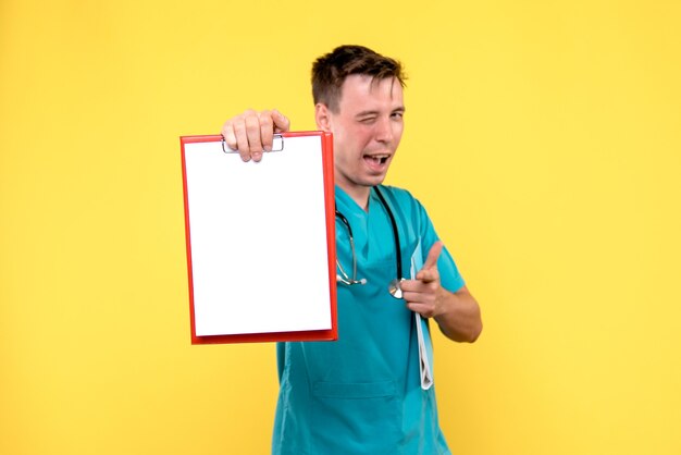 Front view of of young male doctor holding analyzes on yellow wall