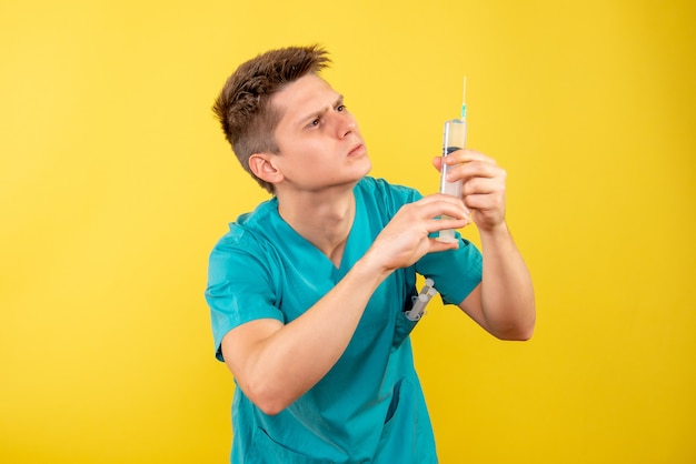 Front view young male doctor in medical suit holding injection on yellow background
