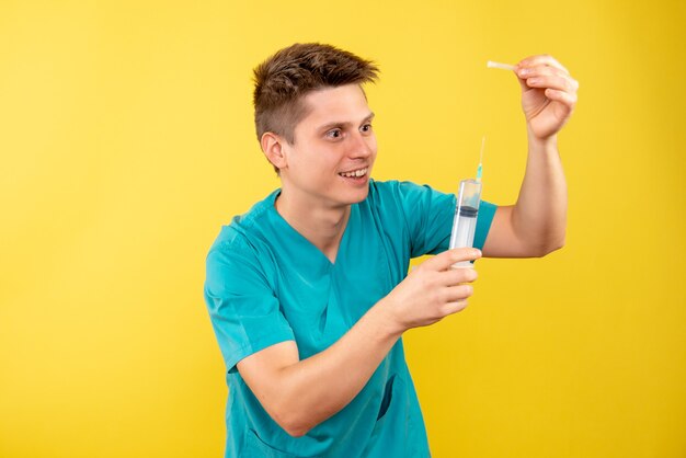 Front view young male doctor in medical suit holding injection on yellow background