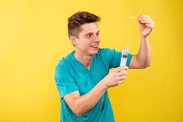 Вид спереди молодой мужчина-врач в медицинском костюме, держащий инъекцию на желтом фоне