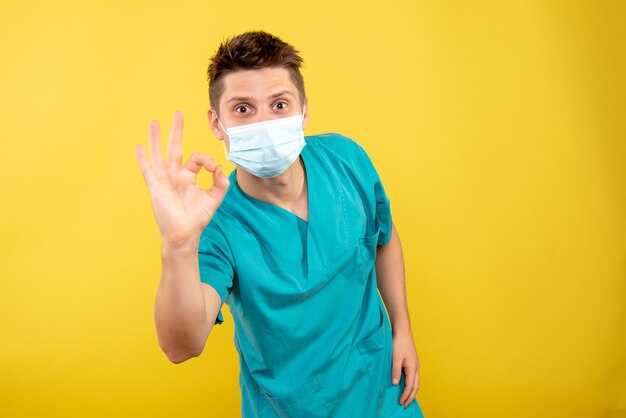 Вид спереди молодой мужчина-врач в медицинском костюме со стерильной маской на желтом фоне