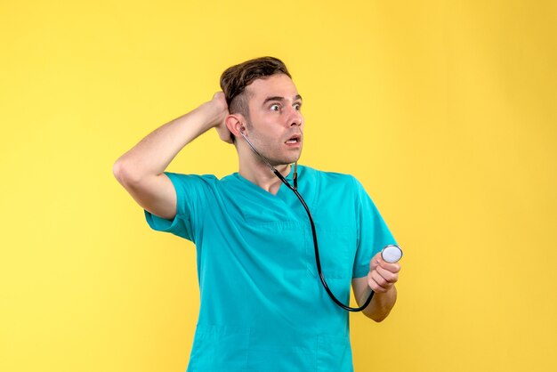 Front view of young male doctor using stethoscope on a yellow wall