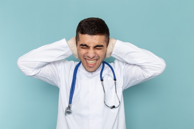 Вид спереди молодого мужчины-врача в белом костюме с синим стетоскопом, закрывающим уши