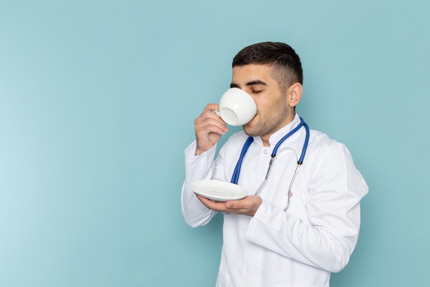 Front view of young male doctor in white suit with blue stethoscope drinking coffee