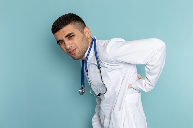 Front view of young male doctor in white suit with blue stethoscope having backache