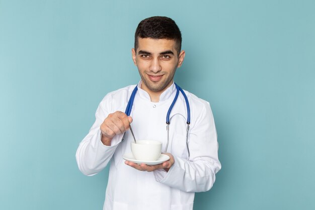 Front view of young male doctor in white suit with blue stethoscope holding cup of coffee and smiling