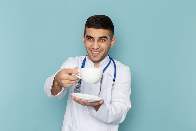 Front view of young male doctor in white suit with blue stethoscope holding cup of coffee with smile
