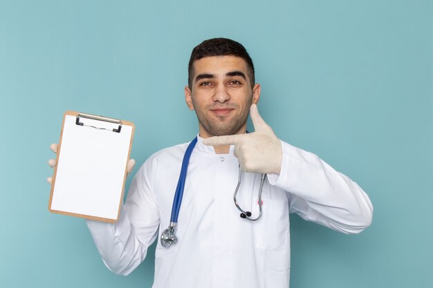 Front view of young male doctor in white suit with blue stethoscope holding notepad with smile