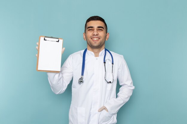 Front view of young male doctor in white suit with blue stethoscope holding notepad