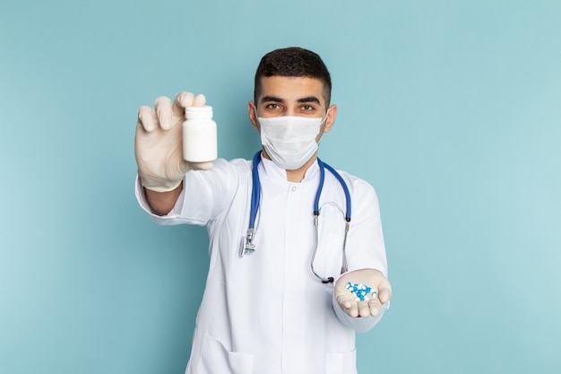 Front view of young male doctor in white suit with blue stethoscope holding pills