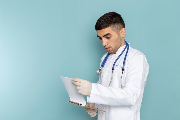 Вид спереди молодого мужчины-врача в белом костюме с синим стетоскопом, держащего и читающего блокнот