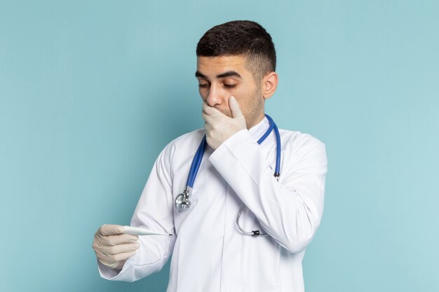 Вид спереди молодого мужчины-врача в белом костюме с синим стетоскопом, держащего термометр с удивленным выражением лица на синем столе, работа в медицинской больнице