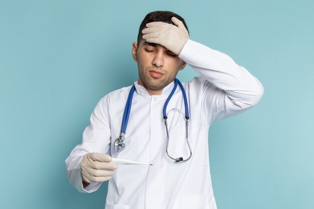 Вид спереди молодого мужчины-врача в белом костюме с синим стетоскопом, держащего термометр