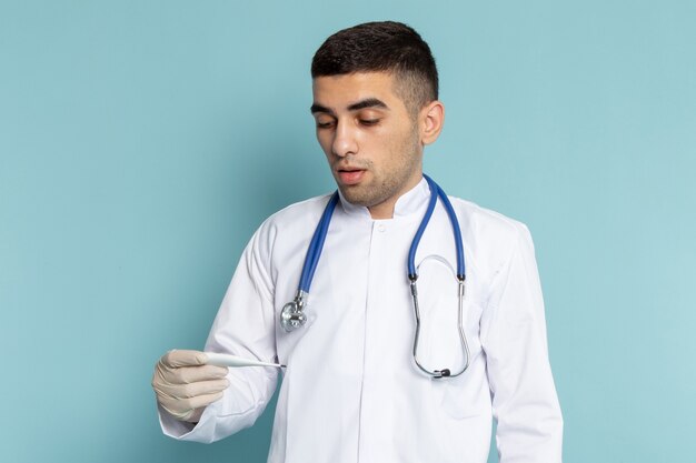 Front view of young male doctor in white suit with blue stethoscope holding thermometer