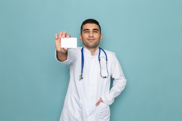 Вид спереди молодого мужчины-врача в белом костюме с синим стетоскопом, держащего белую карточку