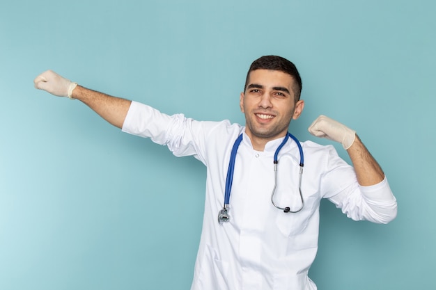 Вид спереди молодого мужчины-врача в белом костюме с улыбкой синего стетоскопа и позирования