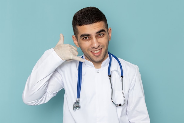 Front view of young male doctor in white suit with blue stethoscope smiling and doing phone sign