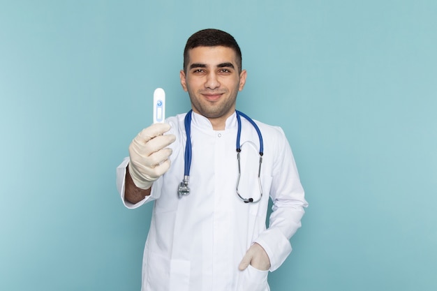 Вид спереди молодого мужчины-врача в белом костюме с синим стетоскопом, улыбающегося и держащего устройство