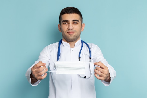 Front view of young male doctor in white suit with blue stethoscope smiling and holding mask