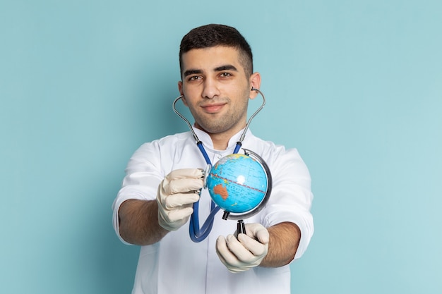 Vista frontale del giovane medico maschio in vestito bianco con lo stetoscopio blu che sorride e che tiene il globo del mondo