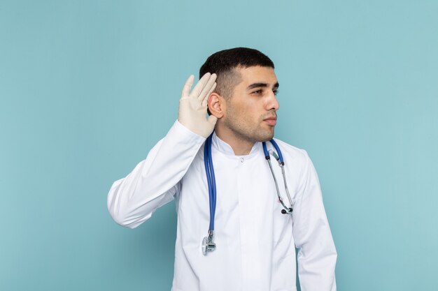 Front view of young male doctor in white suit with blue stethoscope trying to hear out
