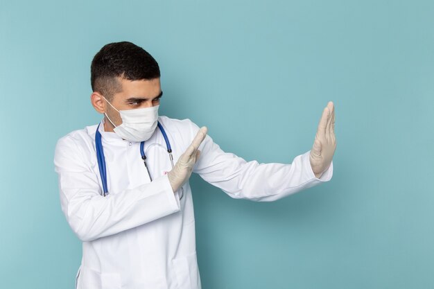 Front view of young male doctor in white suit with blue stethoscope wearing sterile mask posing