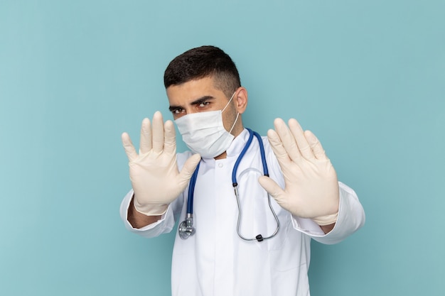 Вид спереди молодого мужчины-врача в белом костюме с синим стетоскопом в стерильной маске с осторожностью