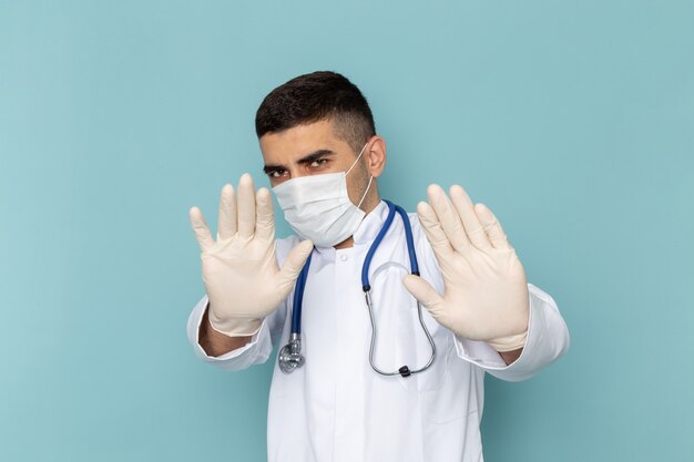 Front view of young male doctor in white suit with blue stethoscope wearing sterile mask with cautious