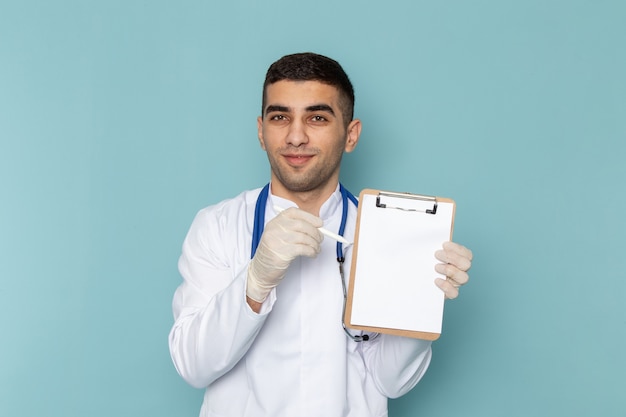 Вид спереди молодого мужчины-врача в белом костюме с синим стетоскопом, записывающим заметки