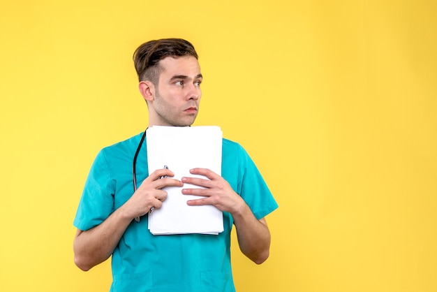 Front view of young male doctor with documents on light yellow wall
