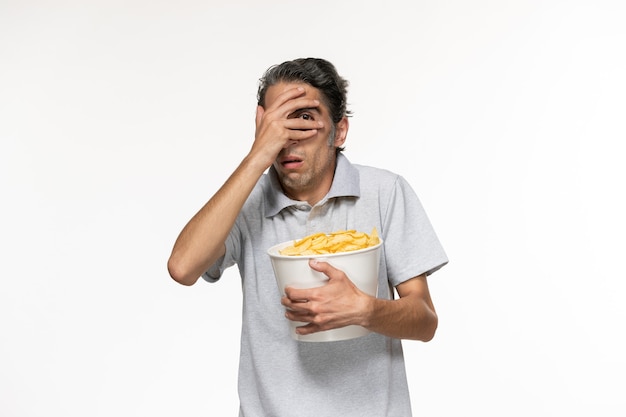 Вид спереди молодой мужчина ест картофельные чипсы, смотрит фильм на светлой белой поверхности