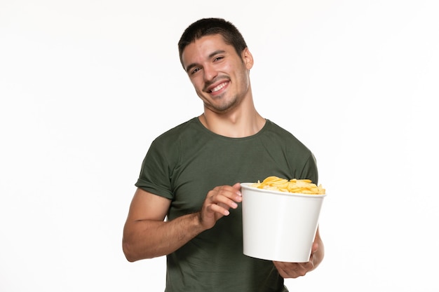 Вид спереди молодой мужчина в зеленой футболке держит корзину с картофельными чипсами и улыбается на белой стене.