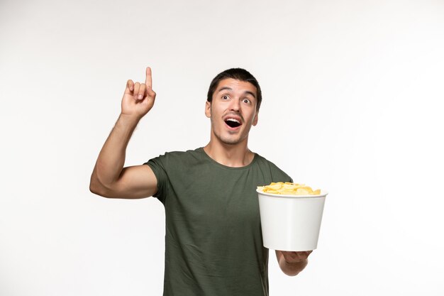 Вид спереди молодой мужчина в зеленой футболке, держащий картофельные чипсы на светлой белой стене, одинокий человек в кино, кино