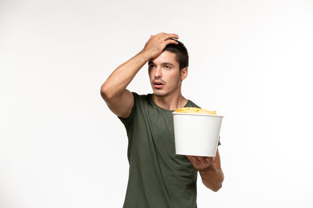 Вид спереди молодой мужчина в зеленой футболке, держащий картофельные чипсы на светлой белой стене, одинокий человек в кино, кино