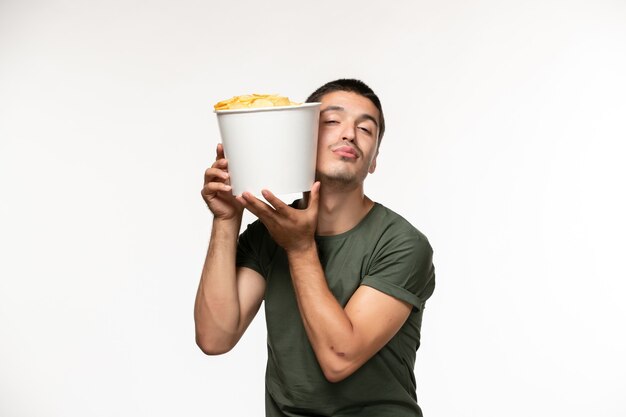 Вид спереди молодой мужчина в зеленой футболке, держащий картофельные чипсы на белой стене, человек одинокий фильм, кино