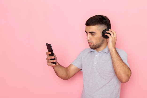 Front view young male in grey shirt holding phone and listening to music with black earphones on pink