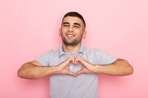 Front view young male in grey shirt smiling and showing love sign on pink