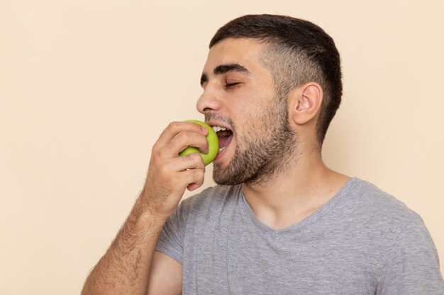 Front view young male in grey t-shirt and blue jeans biting apple on beige